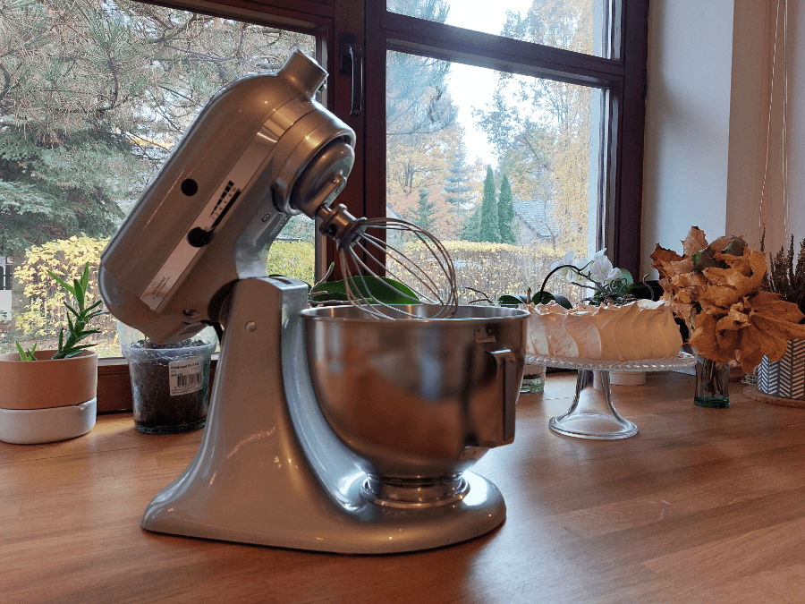 Robot planetarny KitchenAid w kolorze chromowany metal, stoi na drewnianym blacie kuchennym, ma odchyloną głowicę i zamontowane mieszadło - rózgę