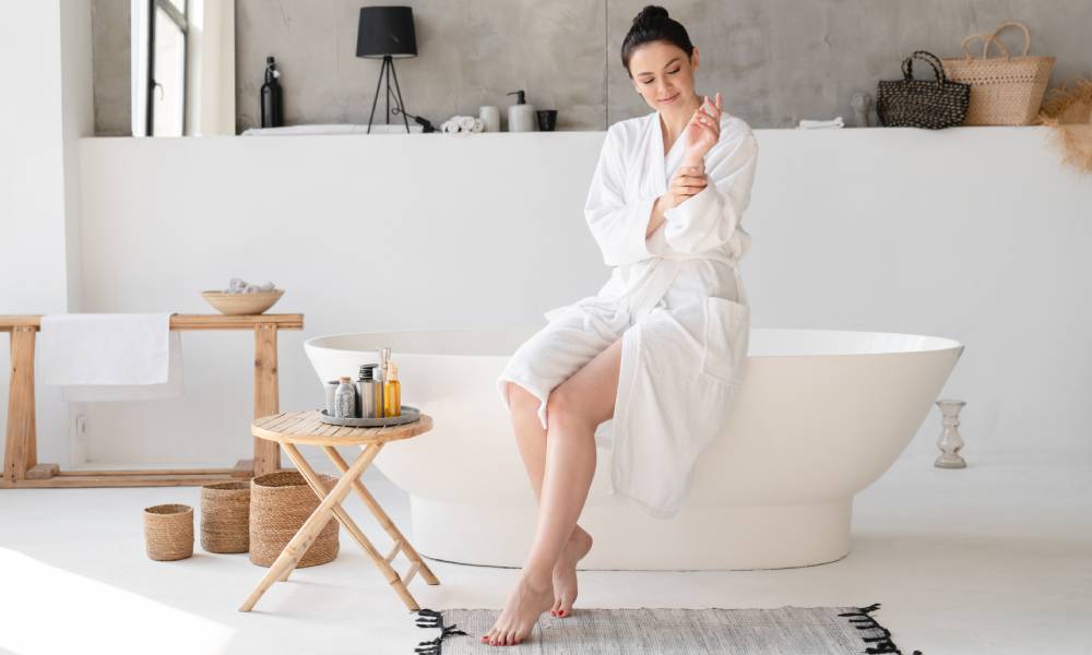 Kobieta siedzi na wannie w nowoczesnej, eleganckiej łazience, jest w trakcie pielęgnacji ciała, zrobiła depilację nóg.