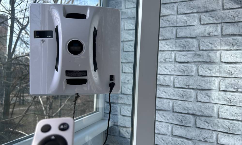 Robot do mycia okien o kwadratowym kształcie pracuje na szybie, a ktoś steruje nim pilotem