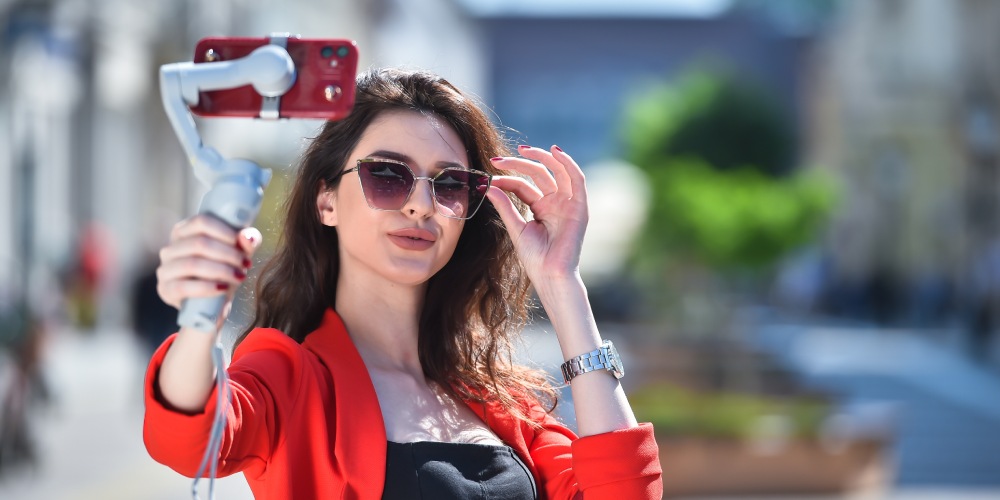 dziewczyna w czerwonej marynarce ma okulary przeciwsłoneczne i trzyma w dłoni gimbala DJI OSMO z telefonem iPhone 12