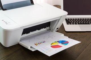  Biała drukarka z kolorowym wydrukiem leży na drewnianym stole, w tle stoi biały laptop