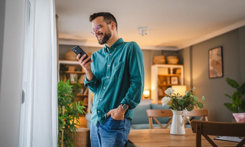 Wesoły mężczyzna steruje na telefonie inteligentnymi funkcjami systemu smart home.