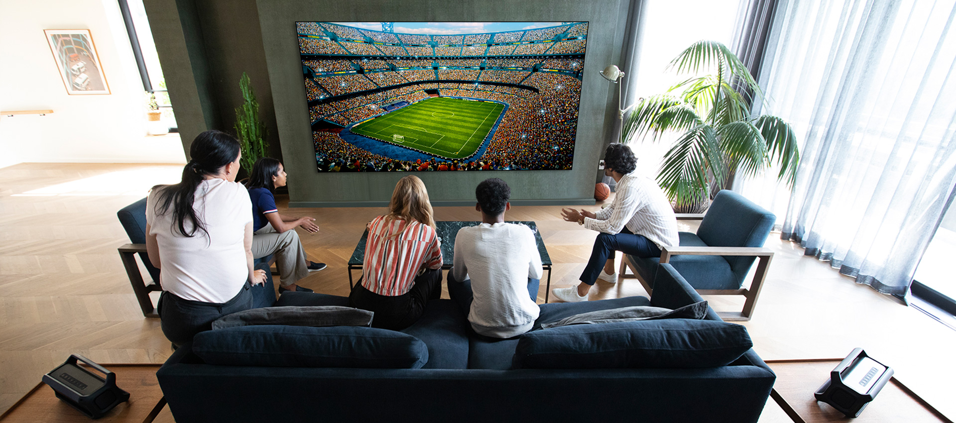 Grupa przyjaciół oglądająca sport w telewizji w pokoju dziennym z dwoma takimi samymi głośnikami tylnymi Bluetooth