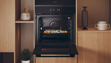 Wbudowany czarny piekarnik Haier z otwartymi drzwiami, w środku taca z warzywami. Piekarnik umieszczony w drewnianej szafce, obok znajduje się półka z białym czajnikiem po lewej stronie oraz przezroczysty brązowy wazon obok drugiego białego czajnika po prawej. Na podłodze przed szafką rośnie mała zielona roślina.
