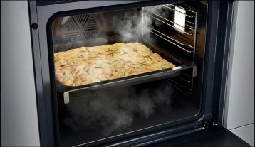 Zbliżenie na piekarnik z otwartymi drzwiami, w środku taca z gorącą focaccią posypaną ziołami, prawdopodobnie rozmarynem. Widać parę, co sugeruje, że jedzenie jest świeżo upieczone. Wnętrze piekarnika ukazuje system półek i oświetlenie wewnętrzne.