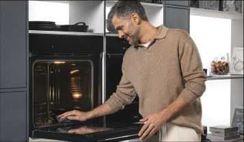 Mężczyzna w beżowym swetrze z krótkimi siwiejącymi włosami sprawdza potrawę w otwartym piekarniku, zintegrowanym w nowoczesnej szafce kuchennej w ciemnoszarym kolorze. Wnętrze piekarnika jest oświetlone, a w środku znajduje się danie. Kuchnia ma jasne ściany i minimalistyczny wystrój, a po prawej stronie widoczna jest półka z dekoracyjnymi przedmiotami i książkami kucharskimi. Mężczyzna uśmiecha się, zadowolony z postępu gotowania.