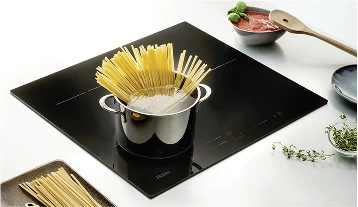 Czarny prostokątny piec kuchenny z stalowym garnkiem wypełnionym makaronem i wodą. Na piecu widoczny jest czerwony wyświetlacz cyfrowy. Obok garnka znajduje się mała taca z dodatkowymi nitkami makaronu, miska z sosem pomidorowym ozdobionym świeżą bazylią oraz drewniana łyżka leżąca na misce. Powierzchnia blatu kuchennego jest biała.