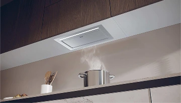 Nowoczesna kuchnia z blatem w marmurowym wzorze w odcieniach bieli, szarości i ciemnego brązu. Na blacie znajduje się stalowy garnek, z którego unosi się para. Nad garnkiem zamontowana jest wbudowana okap kuchenny pod szafką. Szafki kuchenne wykonane są z ciemnego drewna i białego materiału.