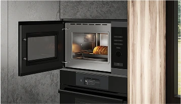 Zbliżenie na mikrofalówkę umieszczoną na górze i piekarnik na dole, oba wbudowane w szafkę kuchenną. Drzwi mikrofalówki są otwarte, a w środku znajduje się częściowo upieczony bochenek chleba. Piekarnik poniżej ma cyfrowy interfejs z małym wyświetlaczem. Styl jest nowoczesny i elegancki, dominują kolory stal nierdzewna i ciemnoszary.