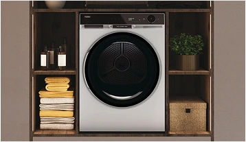 Biała pralka z ciemnymi drzwiami umieszczona w drewnianej jednostce regałowej, z czarnym panelem sterującym nad drzwiami. Po lewej stronie znajdują się starannie złożone kolorowe ręczniki, a po prawej stronie rośnie doniczkowa roślina, pod którą stoi pleciona skrzynka na przechowywanie.