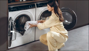 Młoda kobieta w żółtym stroju wkłada ubrania do białej pralki, obok której znajduje się suszarka, w szarym pomieszczeniu.