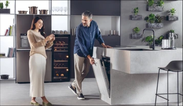 Nowoczesna kuchnia z dobrze oświetlonym wnętrzem, w której para wchodzi w interakcję. Kobieta z długimi, ciemnymi włosami, uśmiechnięta, trzyma telefon, ubrana w beżowy sweter i długą białą spódnicę. Obok niej znajduje się duża chłodziarka na wino. Mężczyzna z siwiejącymi włosami otwiera drzwi do jasnej wbudowanej zmywarki, ubrany w ciemnoniebieską koszulę i ciemne spodnie. W tle widoczna stalowa lodówka oraz różne pojemniki kuchenne. Wyspa kuchenna w jasnym kolorze z ciemnym zlewem, a za zmywarką rosną zielone rośliny w doniczkach. Obok wyspy stoi czarny stołek barowy.