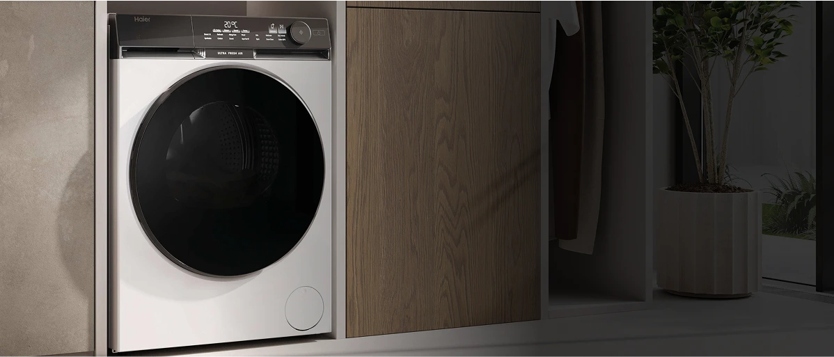 Nowoczesna pralka Haier z białą obudową i czarnymi drzwiami, z panelem sterującym na górze, obok drewniana szafka i roślina w doniczce przy szklanych drzwiach.