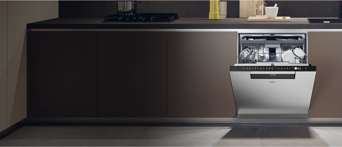 Nowoczesna kuchnia z wbudowaną zmywarką w zabudowie, z gładkimi, bezuchwytowymi szafkami w odcieniach brązu. Otwarta zmywarka w kolorze srebrnym z czarnym panelem sterującym, wypełniona szklankami i talerzami. Na ciemnoszarym blacie znajduje się płyta grzewcza po lewej stronie i zlew po prawej, a obok leży żółty pojemnik i zestaw białych misek. Podłoga wyłożona jasnoszarymi płytkami.