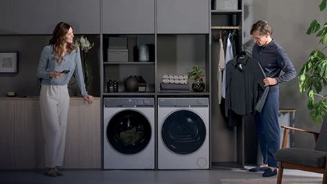 Kobieta i mężczyzna w pralni z pralko–suszarkami Haier w zabudowie. Mężczyzna trzyma koszulę, kobieta opiera się o blat.