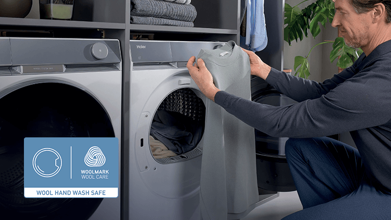 Mężczyzna wyjmuje szary sweter z pralko–suszarki Haier. Obok widoczna etykieta Woolmark potwierdzająca bezpieczne pranie wełny.
