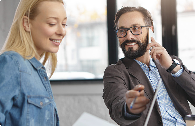 Uśmiechnięty biznesmen w szarym garniturze i krawacie siedzi przy stole i słucha uważnie kobiety siedzącej naprzeciwko niego. Na stole stoją dwie szklanki z wodą.