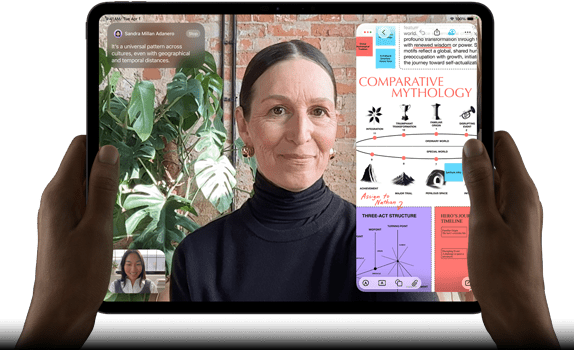 Widoczny od przodu ustawiony poziomo iPad Pro w kolorze gwiezdnej czerni, zaokrąglone rogi, czarna ramka wyświetlacza. Ręce trzymają urządzenie z lewej i prawej strony, na ekranie tłumaczona na żywo rozmowa przez FaceTime. Widać też aplikację edukacyjną z prezentacją „Comparative Mythology”, z tekstem, diagramami i odręcznymi notatkami