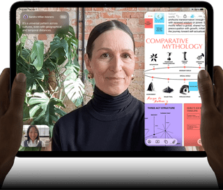 Widoczny od przodu ustawiony poziomo iPad Pro w kolorze gwiezdnej czerni, zaokrąglone rogi, czarna ramka wyświetlacza. Ręce trzymają urządzenie z lewej i prawej strony, na ekranie tłumaczona na żywo rozmowa przez FaceTime. Widać też aplikację edukacyjną z prezentacją „Comparative Mythology”, z tekstem, diagramami i odręcznymi notatkami