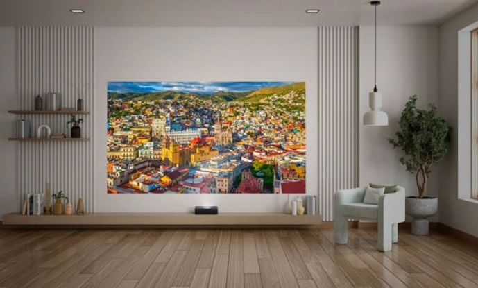 Nowoczesny salon z drewnianymi podłogami, minimalnymi meblami i dużym kolorowym krajobrazem miasta wyświetlanym na ścianie. Obok projekcji znajdują się półki, roślina doniczkowa i biały fotel.