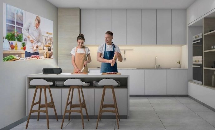 Mężczyzna i kobieta w fartuchach przygotowują jedzenie przy wyspie kuchennej. Przy blacie znajdują się trzy stołki, a ekran na ścianie pokazuje szefa kuchni demonstrującego przepis. Kuchnia jest nowoczesna i jasna z białymi szafkami.