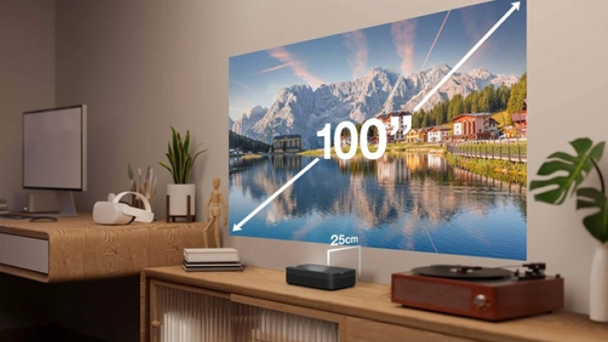 Projektor na drewnianej szafce wyświetla żywy 100-calowy obraz gór i jeziora na ścianie; odległość od projektora do ściany jest oznaczona jako 25 cm. Widoczny jest również gramofon i materiały biurowe.