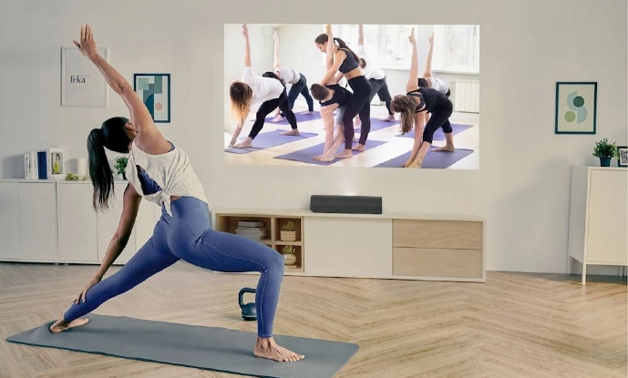 Kobieta ćwiczy jogę na macie w swoim salonie, śledząc zajęcia jogi wyświetlane na dużym ekranie na ścianie. Pokój jest jasny, z minimalistycznym wystrojem i drewnianą podłogą.