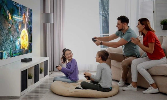 Czteroosobowa rodzina gra razem w grę wideo w swoim salonie. Dwoje dzieci siedzi na podłodze z kontrolerami, podczas gdy rodzice, siedzący na kanapie, grają i kibicują. Na telewizorze wyświetlana jest kolorowa gra.