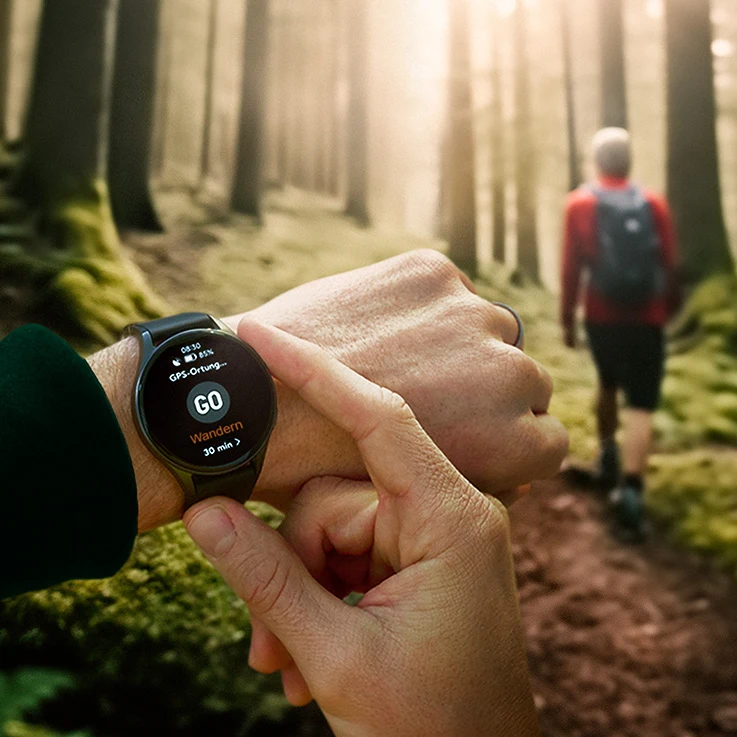 Zdjęcie mężczyzny z zegarkiem na ręku, w tle osoba wedrująca w lesie