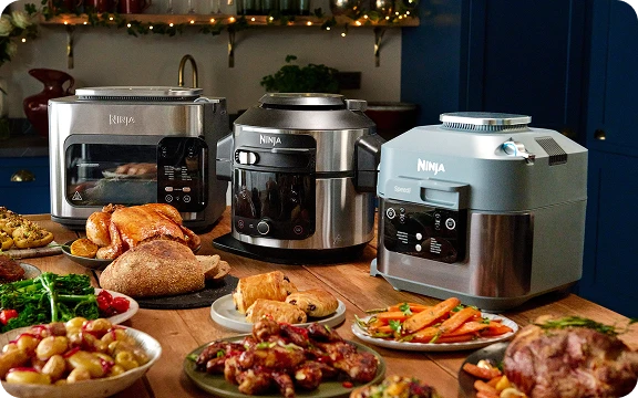 Trzy urządzenia kuchenne Ninja (toster, multicooker i szybkowar) otoczone bogato zastawionym stołem z jedzeniem