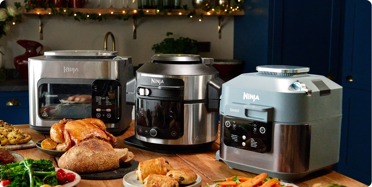 Trzy urządzenia kuchenne Ninja (toster, multicooker i szybkowar) otoczone bogato zastawionym stołem z jedzeniem