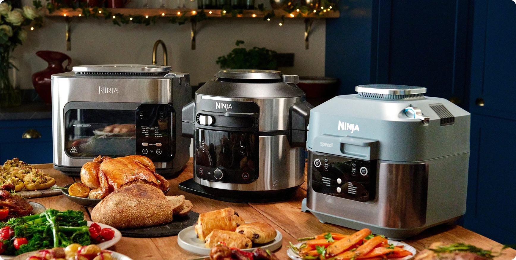 Trzy urządzenia kuchenne Ninja (toster, multicooker i szybkowar) otoczone bogato zastawionym stołem z jedzeniem