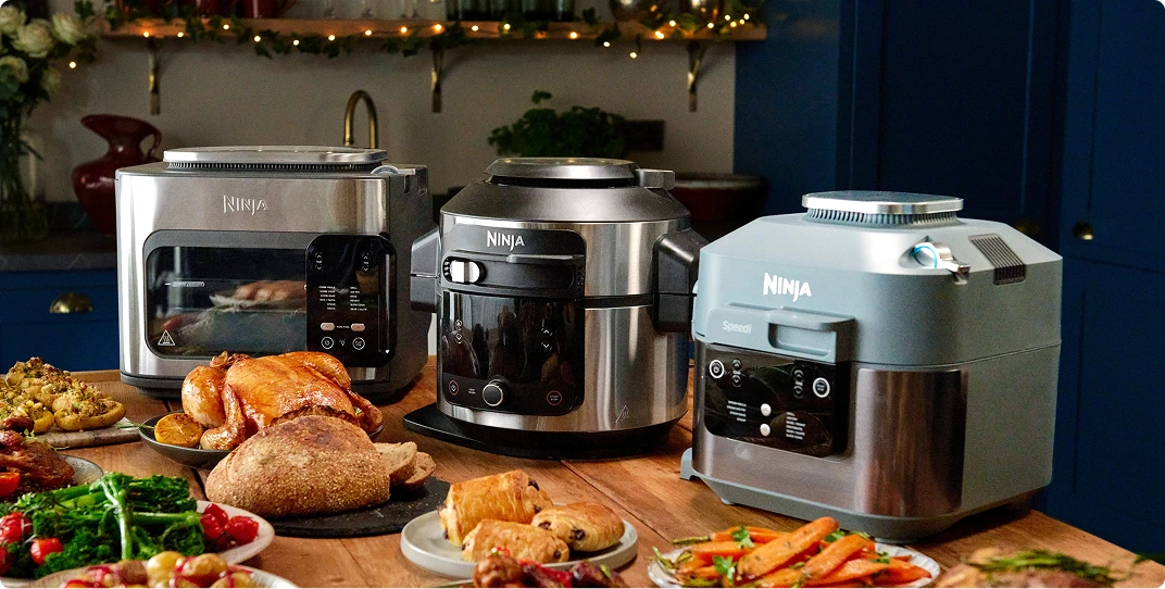 Trzy urządzenia kuchenne Ninja (toster, multicooker i szybkowar) otoczone bogato zastawionym stołem z jedzeniem