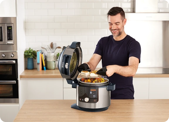 Uśmiechnięty mężczyzna wyjmuje rybę i warzywa z otwartego multicookera Ninja w nowoczesnej, jasnej kuchni