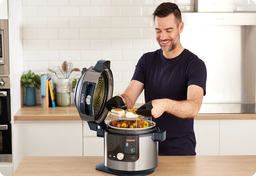 Uśmiechnięty mężczyzna wyjmuje rybę i warzywa z otwartego multicookera Ninja w nowoczesnej, jasnej kuchni