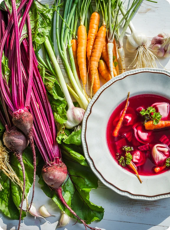 Widok od góry na talerz czerwonego barszczu z uszkami. Obok leżą marchewki, buraki i inne składniki zupy.