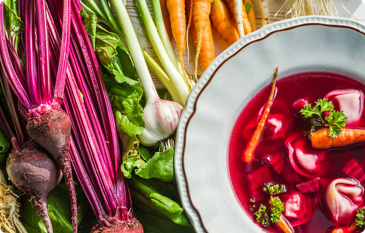 Widok od góry na talerz czerwonego barszczu z uszkami. Obok leżą marchewki, buraki i inne składniki zupy.