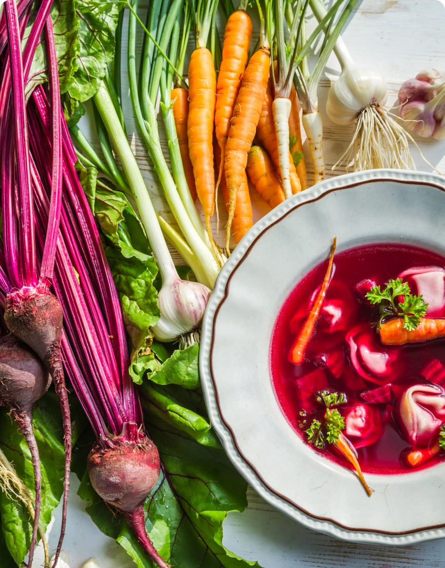 Widok od góry na talerz czerwonego barszczu z uszkami. Obok leżą marchewki, buraki i inne składniki zupy.