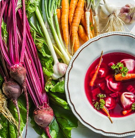 Widok od góry na talerz czerwonego barszczu z uszkami. Obok leżą marchewki, buraki i inne składniki zupy.