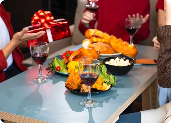 4 osoby na imprezie świątecznej, dzielą się prezentami przy stole, na którym są różne potrawy i lampki wina