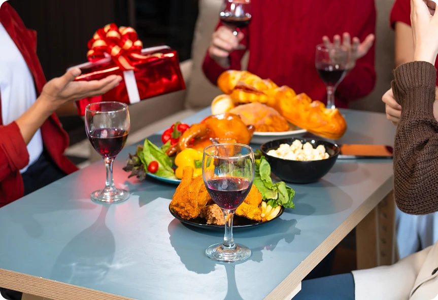 4 osoby na imprezie świątecznej, dzielą się prezentami przy stole, na którym są różne potrawy i lampki wina