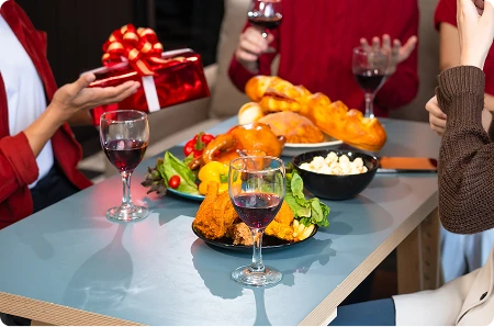 4 osoby na imprezie świątecznej, dzielą się prezentami przy stole, na którym są różne potrawy i lampki wina