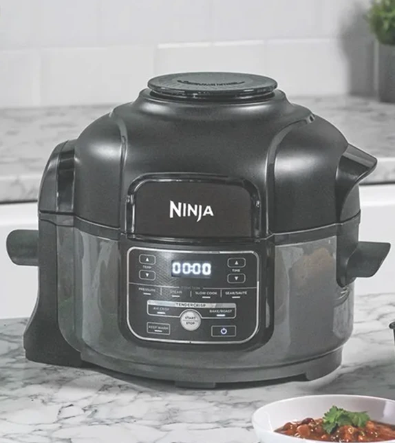 Ninja Foodi MINI OP100EU stoi na blacie kuchennym obok ciasta w foremce i różnych potraw świątecznych