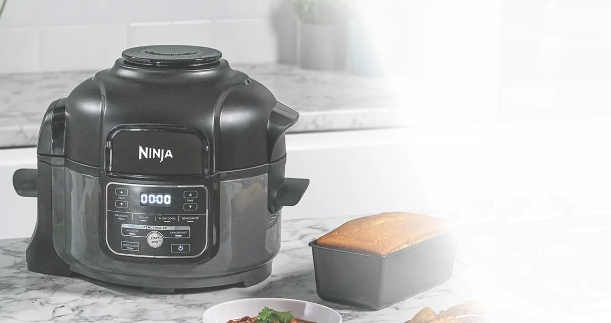 Ninja Foodi MINI OP100EU stoi na blacie kuchennym obok ciasta w foremce i różnych potraw świątecznych