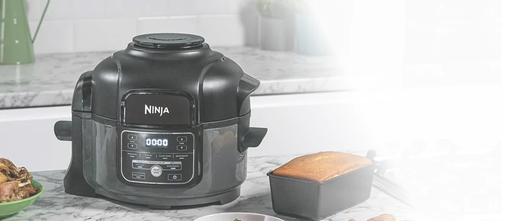 Ninja Foodi MINI OP100EU stoi na blacie kuchennym obok ciasta w foremce i różnych potraw świątecznych