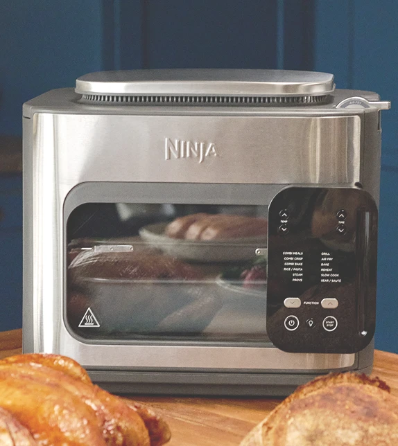 Ninja Combi SFP700EU na stole w kuchni w otoczeniu różnych potraw, w tym kurczaka, pieczonej marchewki i pieczywa