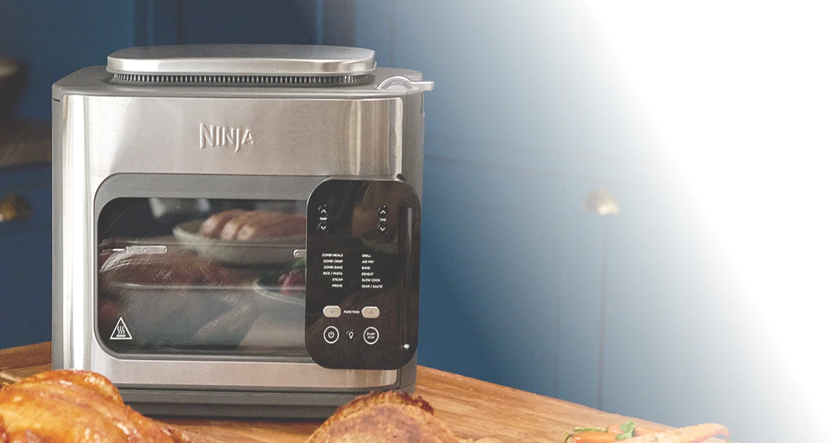 Ninja Combi SFP700EU na stole w kuchni w otoczeniu różnych potraw, w tym kurczaka, pieczonej marchewki i pieczywa