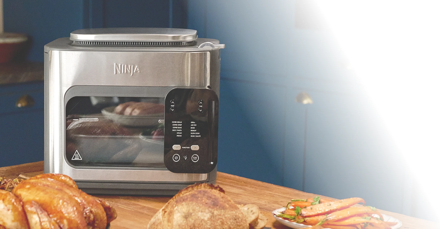 Ninja Combi SFP700EU na stole w kuchni w otoczeniu różnych potraw, w tym kurczaka, pieczonej marchewki i pieczywa