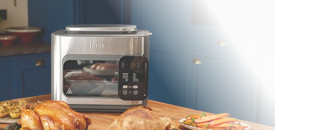 Ninja Combi SFP700EU na stole w kuchni w otoczeniu różnych potraw, w tym kurczaka, pieczonej marchewki i pieczywa