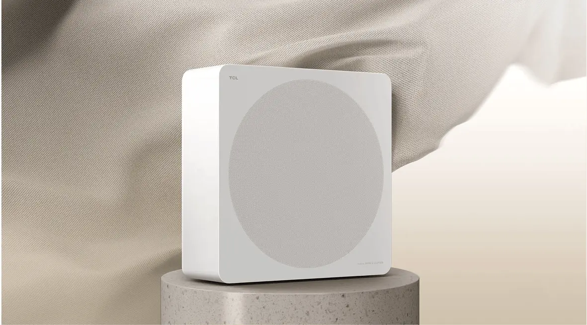 Biały subwoofer TCL Bang & Olufsen z okrągłym głośnikiem umieszczony na betonowej podstawie z jasną tkaniną w tle